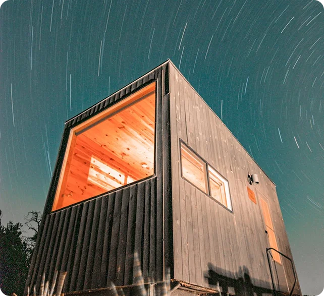 Wooden cabin with glowing windows under night sky with star trails